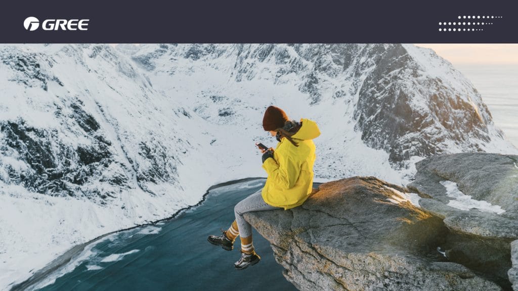 Talvinen vuoristomaisema, jossa vuoren rinteellä istuu keltatakkinen nainen puhelinta katsoen.