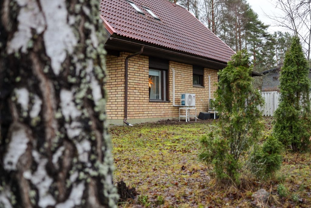 Isohko kaksikerroksinen talo syksyisessä säässä kuvattuna sivulta päin. Kuvassa myös GREE SP Nordic -ilmalämpöpumpun ulkoyksikkö asennettuna maatukitelineelle.